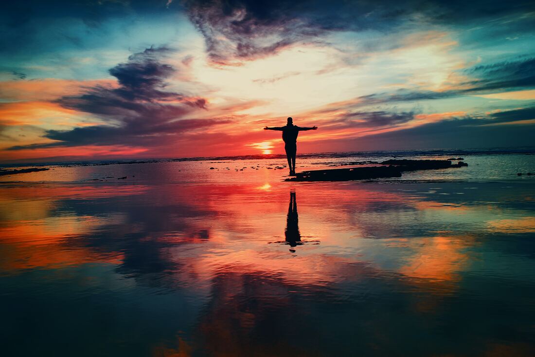 Ihminen näkyy mustana siluettina seisoessaan rannalla taustallaan värikäs auringonlasku. Ihmisellä on kädet levitettyinä sivuilleen.